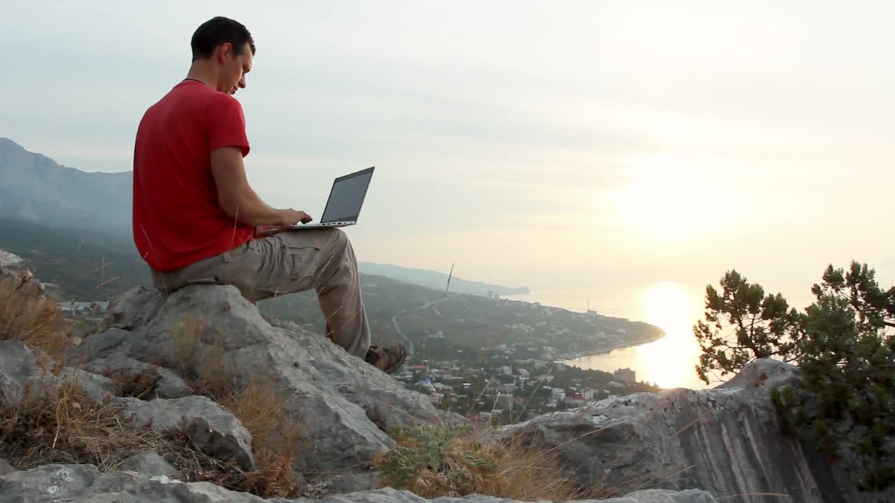 一个年轻人坐在俯瞰城市和大海的悬崖边上，一边欣赏风景优美的日出，一边拿着笔记本电脑远程工作。自由和自由工作的本质视频素材