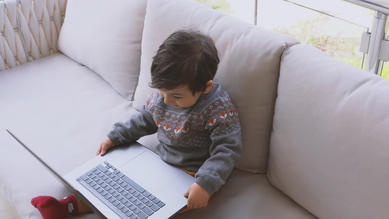 可爱的小男孩坐在阳台的沙发上使用笔记本电脑视频下载