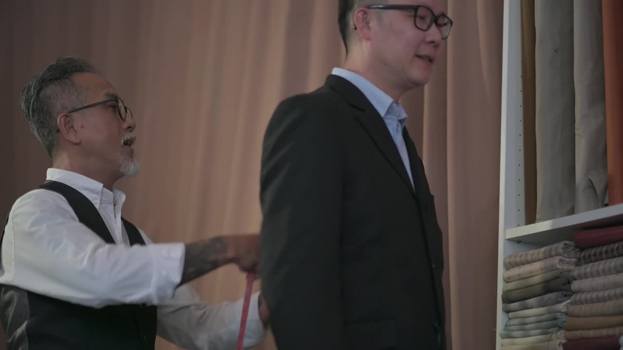 亚洲华人成熟男子裁缝与脸胡茬测量他的客户的身体尺寸，为新的运动上衣制作视频素材