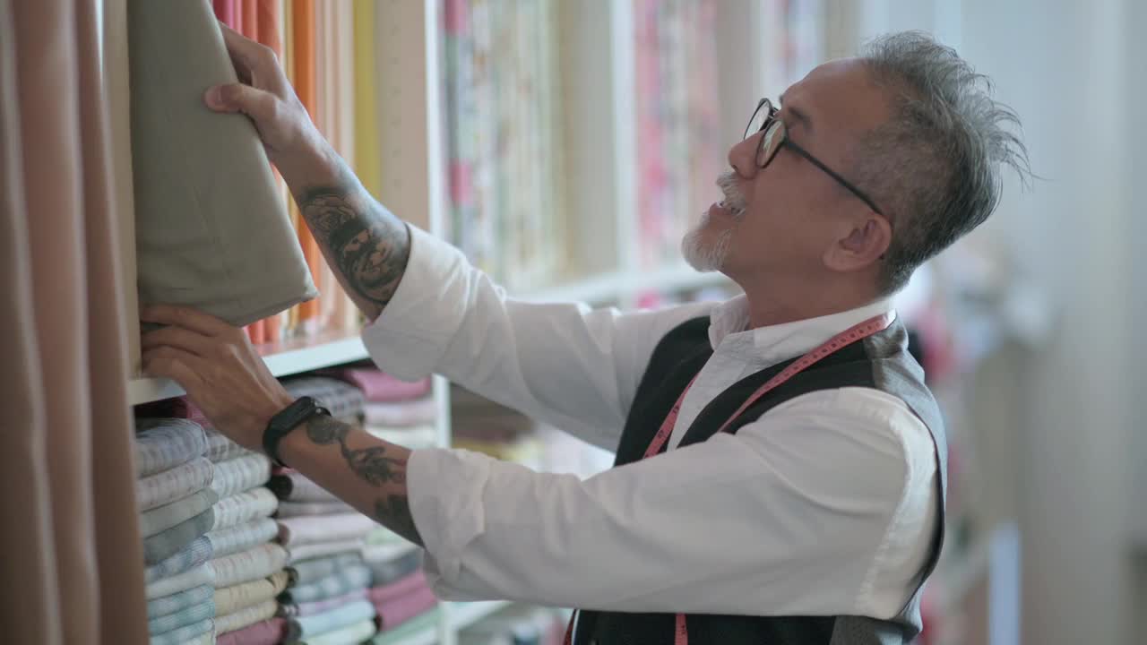 面部留胡茬的亚洲华人成熟男子裁缝在工作室检查他的织物服装视频下载