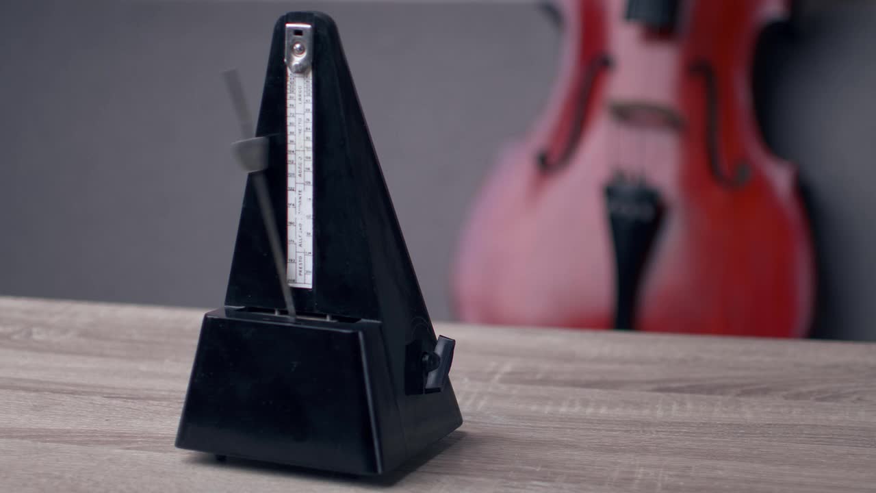 一个黑色复古节拍器站在桌子上敲击着节奏，后面放着一把大提琴，这是一个特写镜头视频下载
