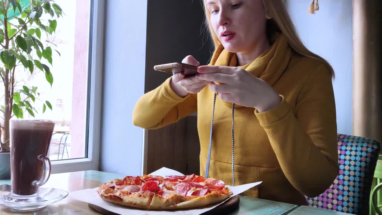 一个博主女孩坐在咖啡馆里用智能手机拍披萨。在社交网络、视频博客和照片博客中工作。创建互联网内容和食物摄影的概念。视频下载