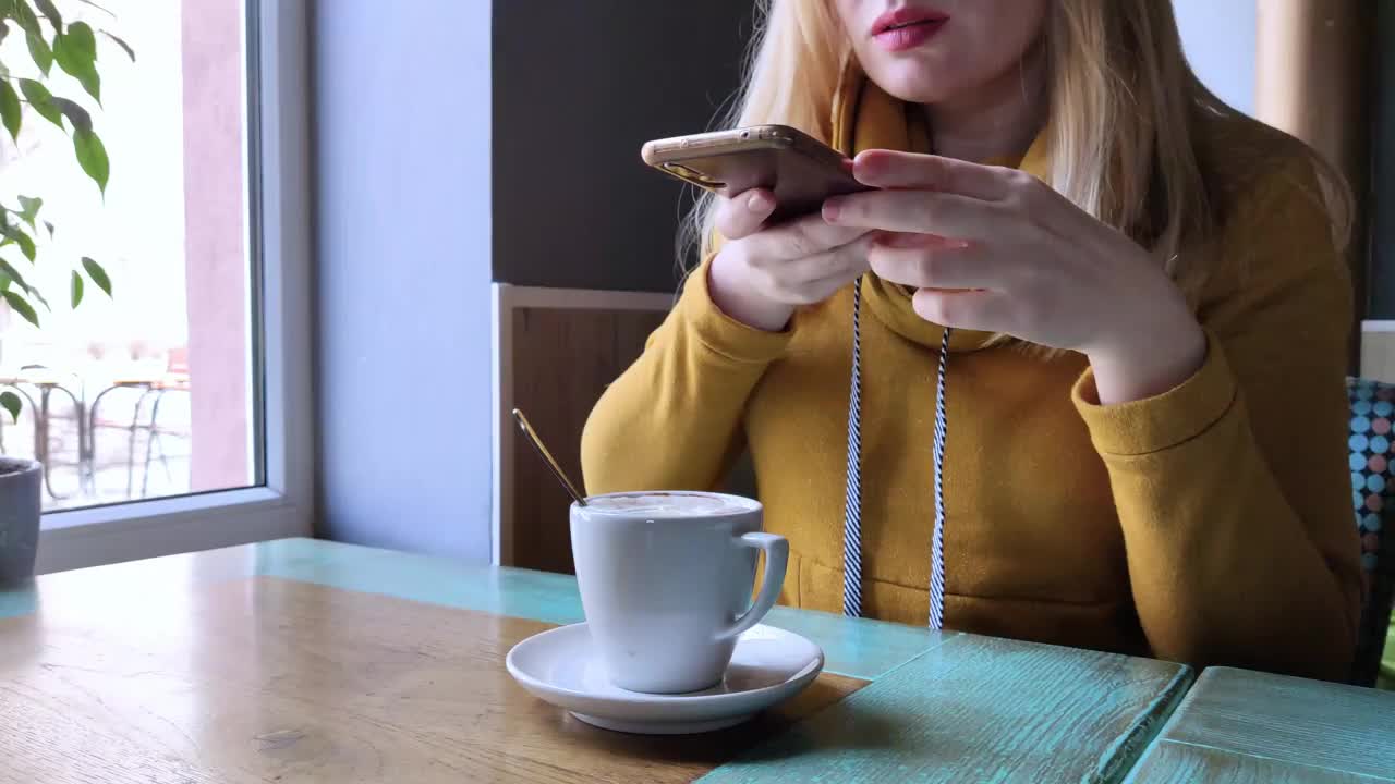 博主女孩坐在咖啡馆里用智能手机拍一杯卡布奇诺泡沫。在社交网络、视频博客和照片博客中工作。互联网内容创作和食物摄影的概念。视频下载
