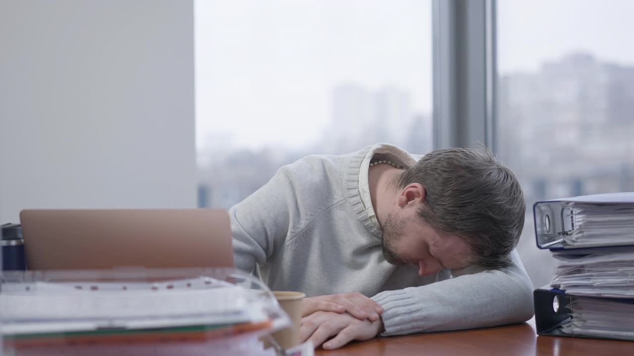 年轻疲惫的黑发男人睡在办公室的桌子上。疲惫的白人男性雇员躺在室内的工作场所。业务倦怠、疲劳和疲惫的概念。视频下载