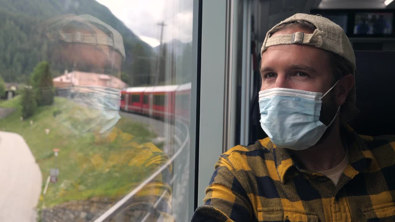 一个戴着外科口罩乘火车旅行的年轻人向窗外望去。视频下载