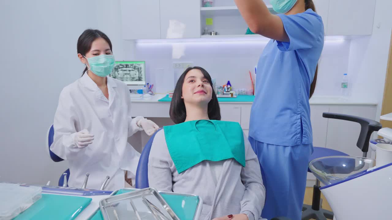 亚洲女牙医调整牙科手术灯，然后开始检查或检查年轻女孩病人的牙齿躺在牙科椅上。牙科助理在牙科诊所提供器械支援。视频下载