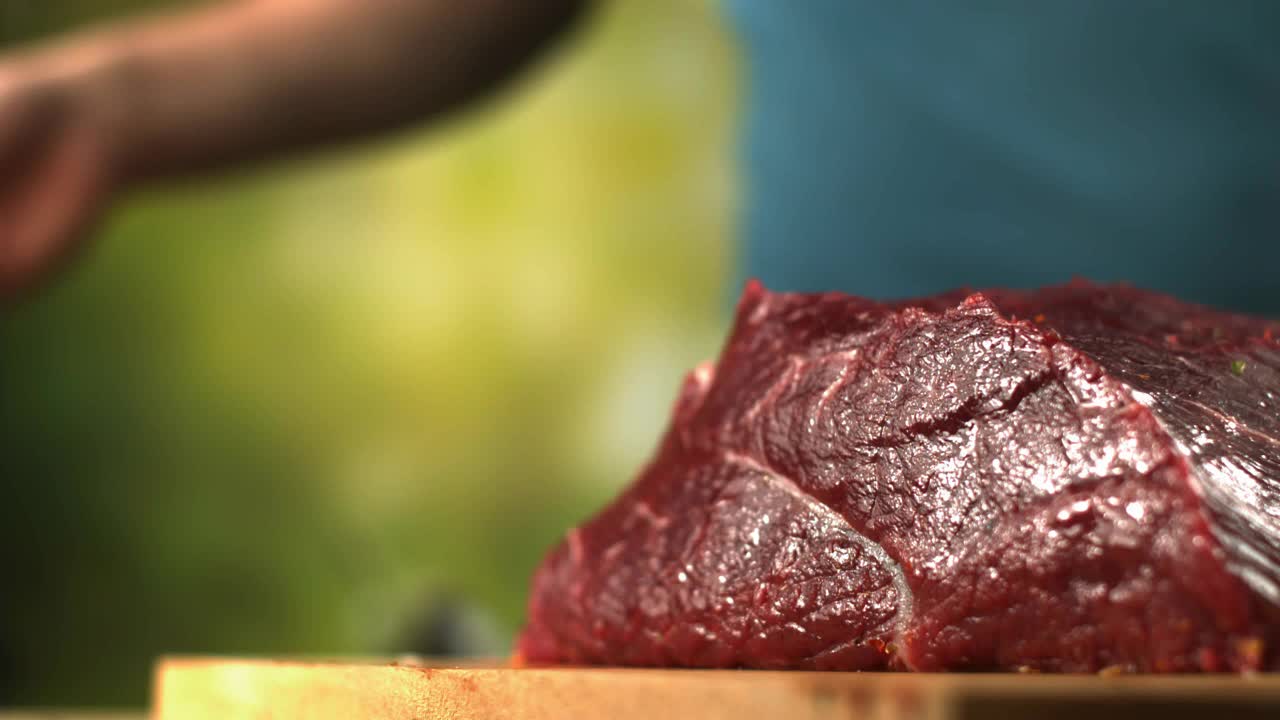 在绿色的背景下，厨师正在用慢动作将胡椒和盐洒在一块木头砧板上的新鲜牛肉上视频下载
