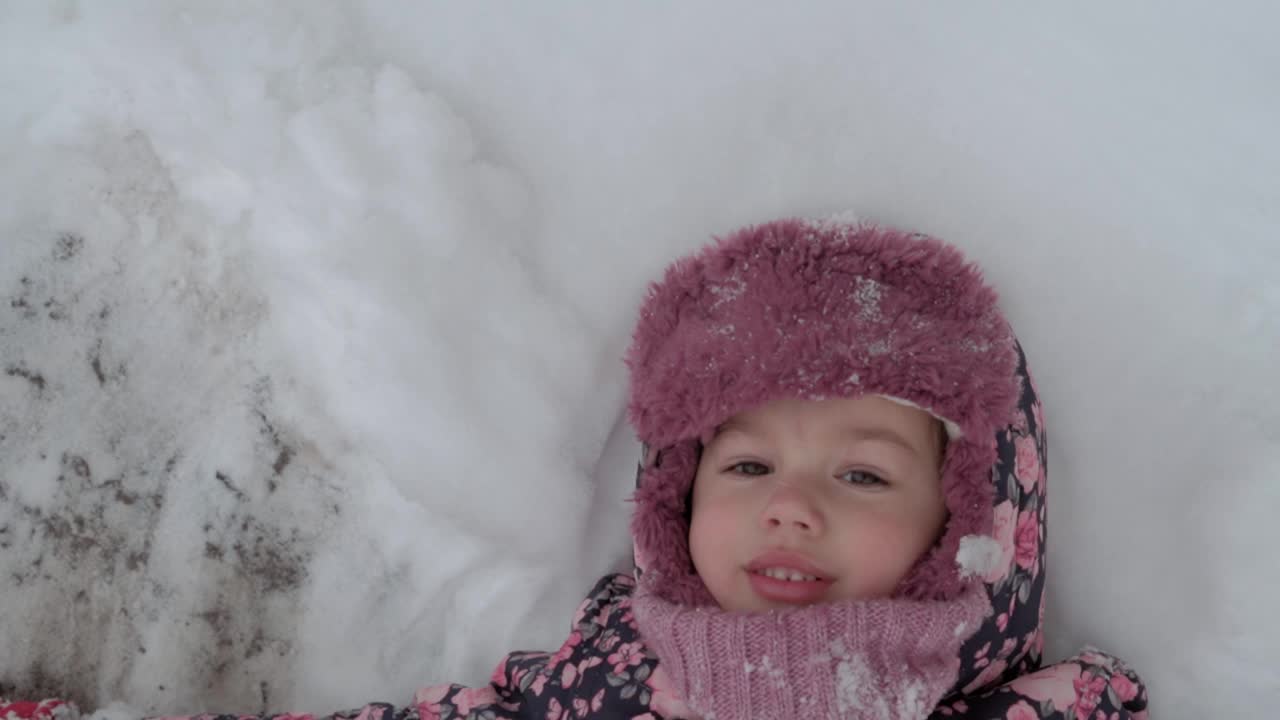 假期，游戏，童年概念慢动作正宗快乐学步的小女孩微笑躺在雪地上，用脚鼓掌做天使。冬季天气寒冷，冬季户外下雪视频下载
