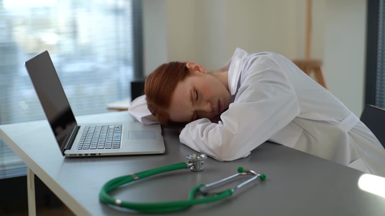 追踪镜头疲惫疲惫过度工作的年轻女医生在办公桌上睡觉的白大褂笔记本电脑和听诊器在工作场所。视频下载