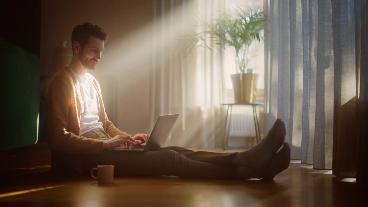 英俊快乐的男人使用笔记本电脑在家远程工作。坐在地板上微笑的家伙使用互联网，写报告，完成项目，使用互联网。远程工作的好晴天视频下载