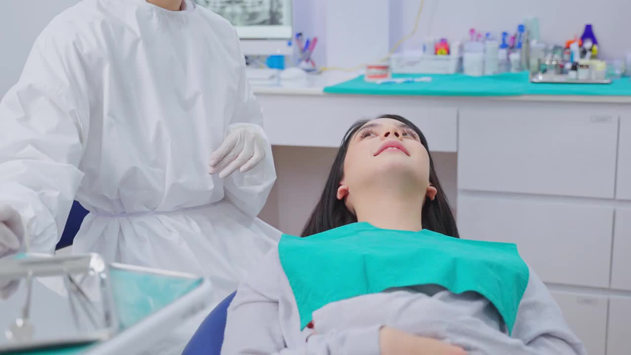 亚洲女牙医穿着实验服，戴着防护面罩和护罩，在诊所治疗过程中为年轻女孩病人在牙科椅上调整牙科手术灯和检查牙齿视频下载