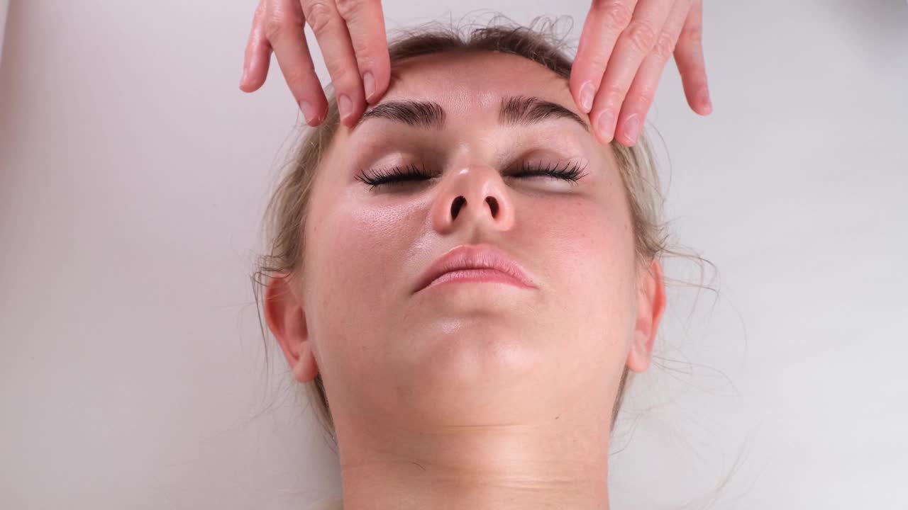 面部按摩美容治疗。一个年轻女子的脸的近距离躺在背上，得到面部提拉按摩，捏和滚动技术视频下载