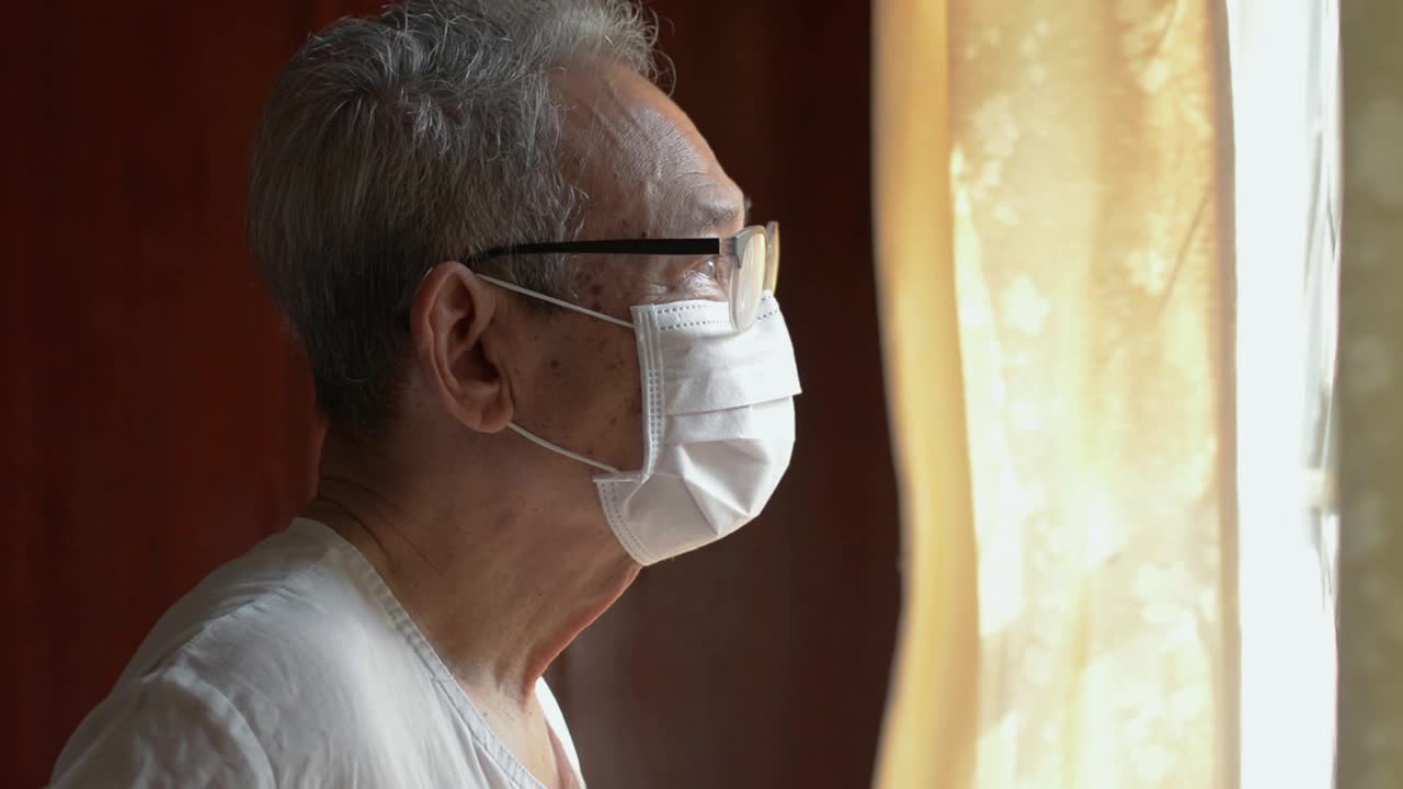 亚洲灰白头发的老年男子戴着口罩戴着眼镜从窗口往外看。视频素材