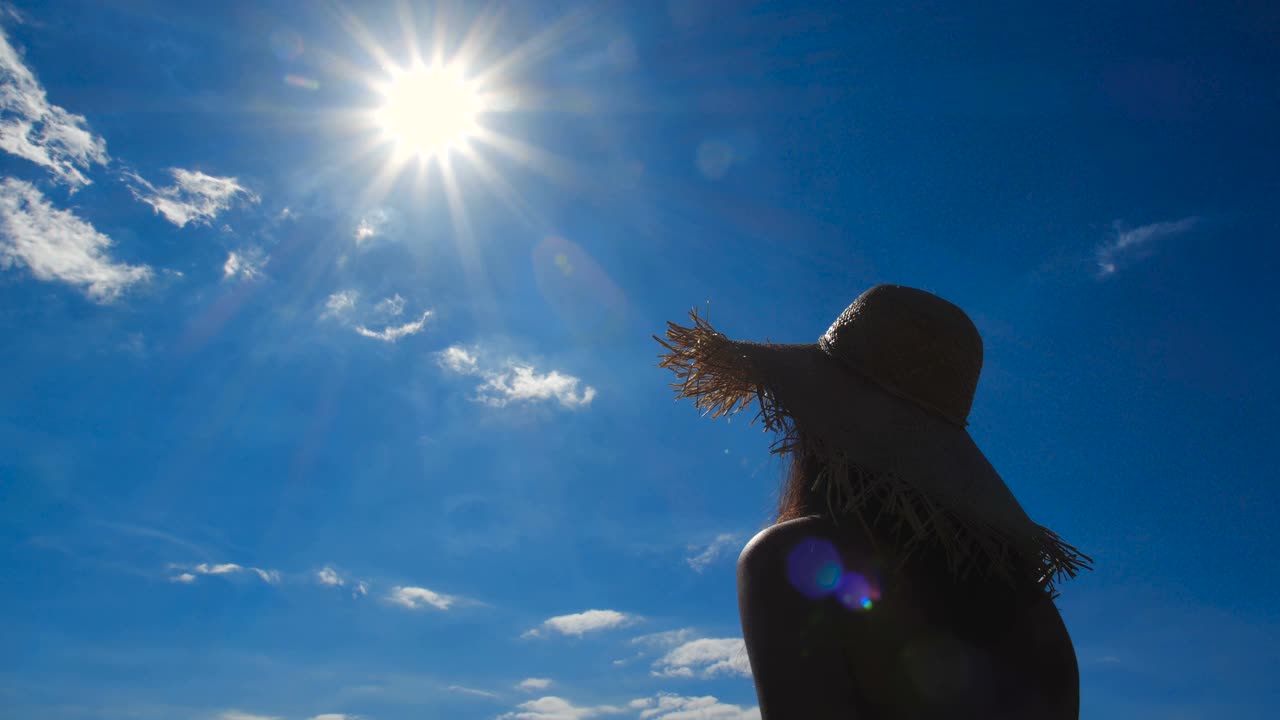 近剪影的美丽的年轻女子在草帽，喝菠萝鸡尾酒和伸出她的手对太阳的背景蓝色的天空。4k 50fps慢动作视频下载