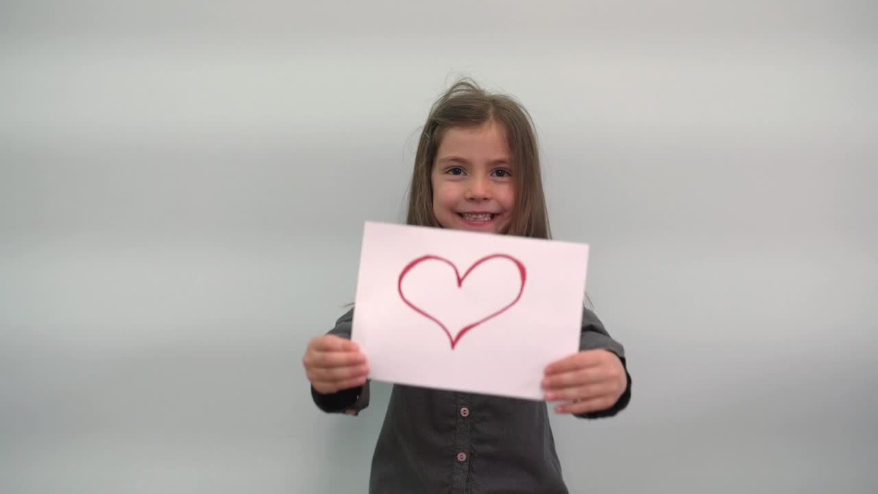 一个女孩拿着一张画着一颗心的纸，微笑着。一个孩子把一个在灰蓝色背景上有一颗心的标志变成了她的手视频下载