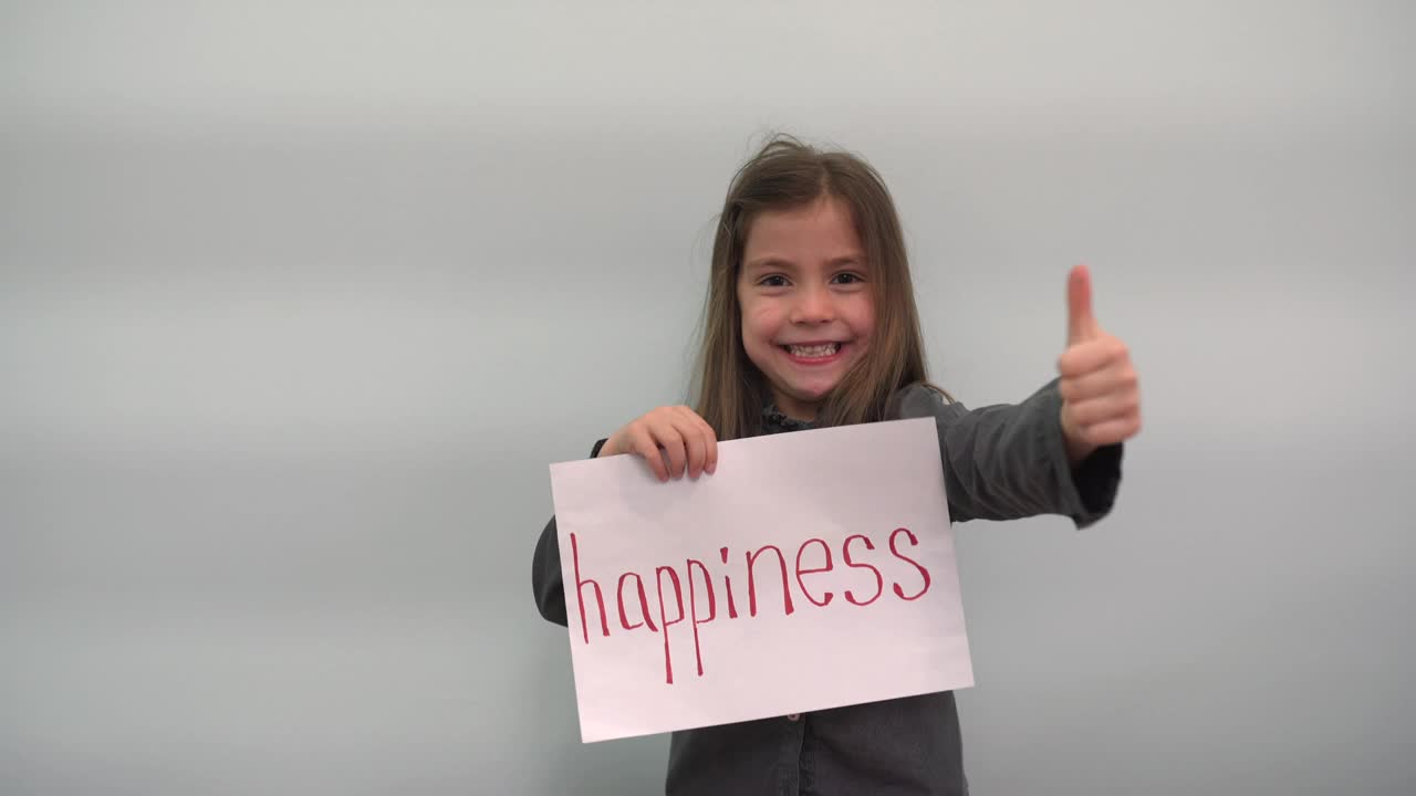 女孩拿着一张写有“幸福”字样的纸，向全班展示并微笑。孩子在他的手中转动着一个灰色蓝色背景上写有“幸福”字样的手势视频下载