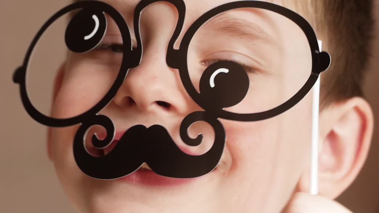 一个快乐，有趣的学龄前儿童拿着胡子和眼镜在他的脸前，孩子描绘一个严格的父母。这是一个孩子关于大人的笑话。父亲节幽默。有选择性的重点,视频下载