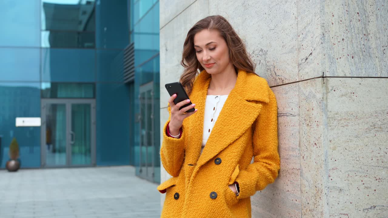 商务女性穿着黄色外套站在户外法人大厦的背景与智能手机打字短信打电话视频下载