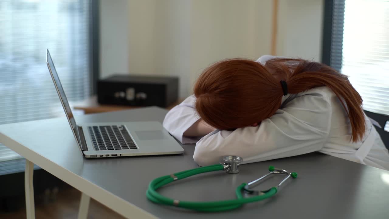 特写劳累过度的红发女医生在白大褂睡觉在书桌上的笔记本电脑和听诊器在工作地点靠近窗户视频下载