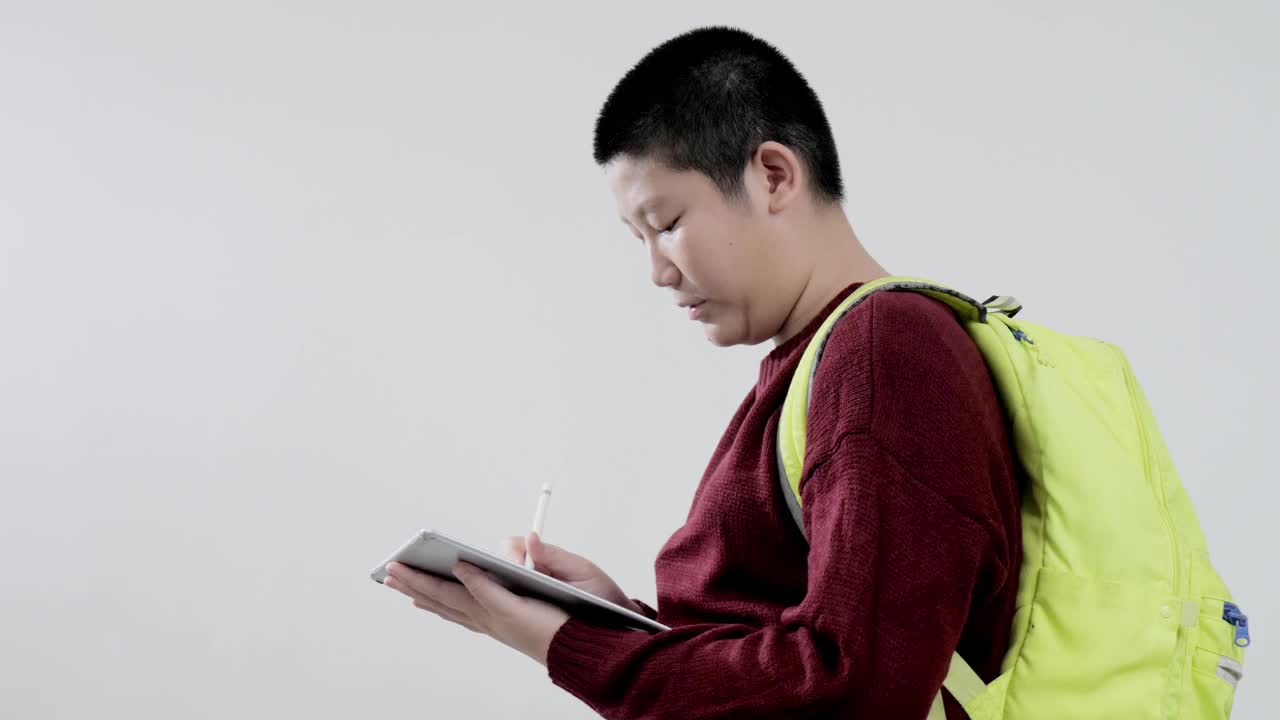 亚洲男生穿着红色毛衣，用数码平板电脑在灰色背景上写笔记，教育生活理念。视频下载