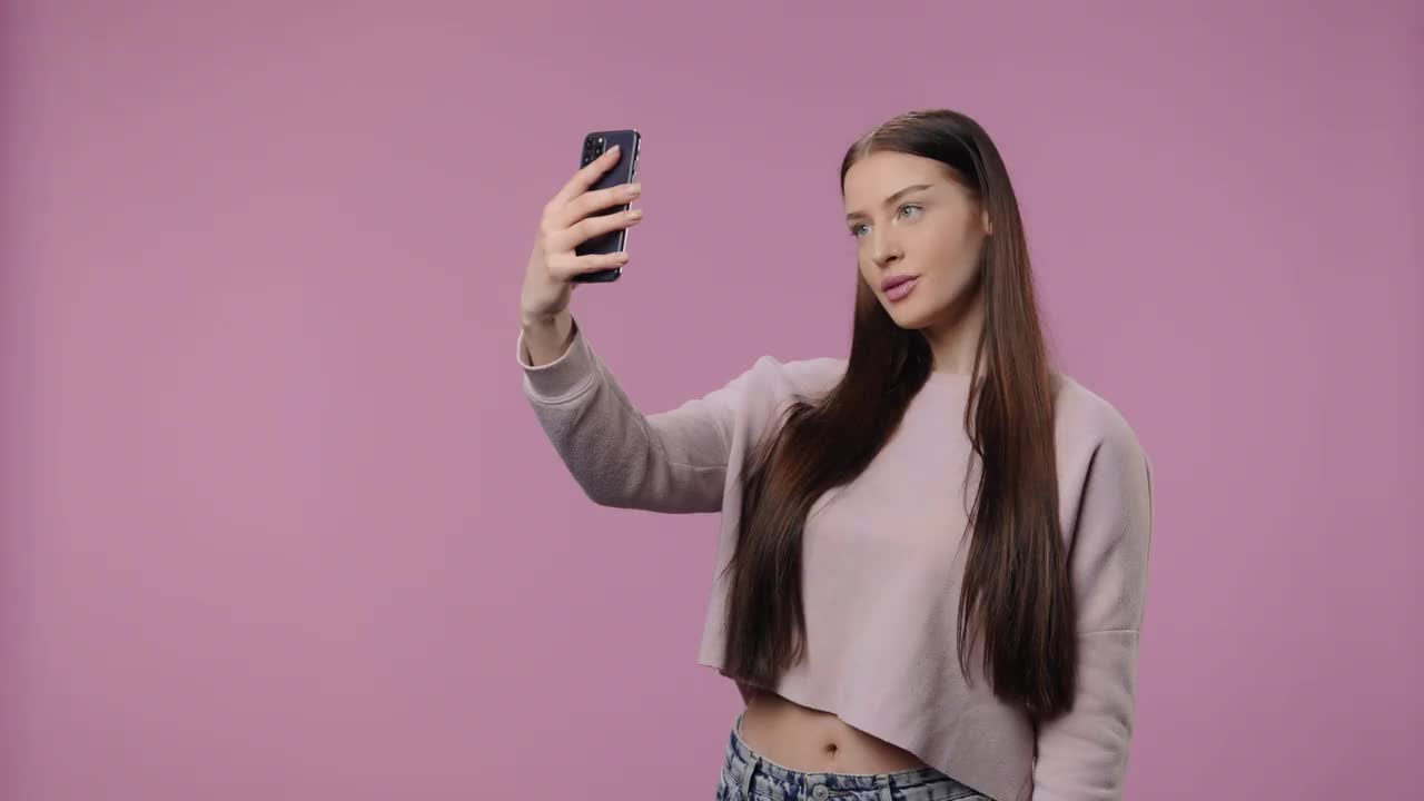一个美容模特在手机上自拍，然后看照片。年轻女孩与长头发在紫色工作室背景上摆姿势。关闭了。慢动作准备59.94fps视频下载