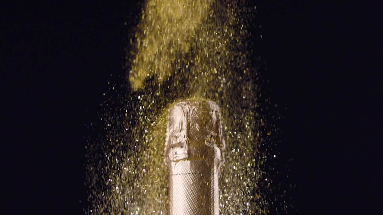 在黑色的背景下，金色的粉末沿着香槟瓶流动。摄像机从上到下。慢镜头，Blackmagic Ursa Pro G2, 300 fps。视频下载