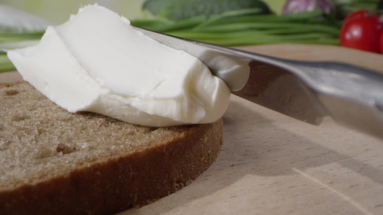 用黄油刀把奶酪涂在烤黑麦面包上，在厨房的桌子上，背景是蔬菜视频下载