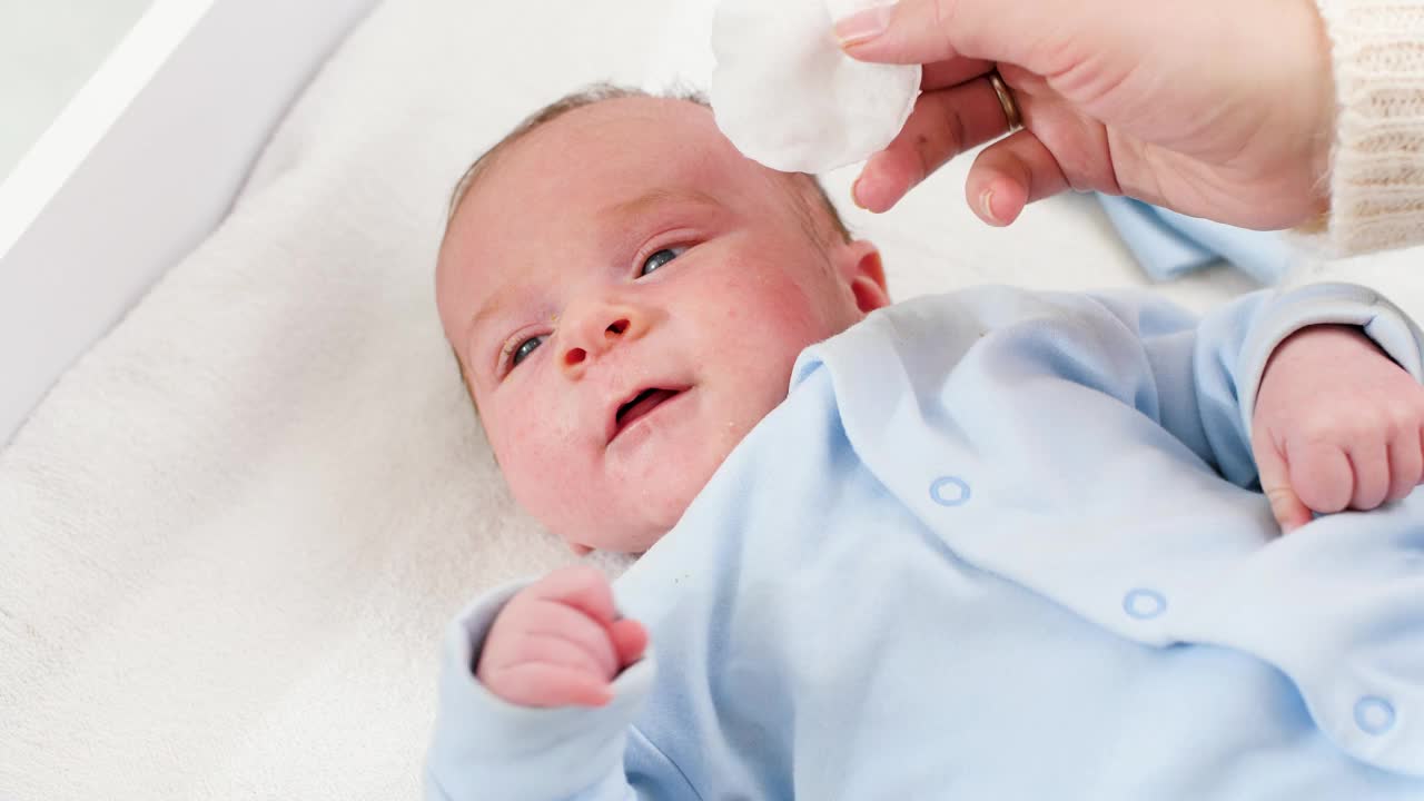 母亲用棉垫清洁新生儿的脸和眼睛的特写。婴儿和新生儿卫生保健概念。有小孩的有爱心的父母。视频下载
