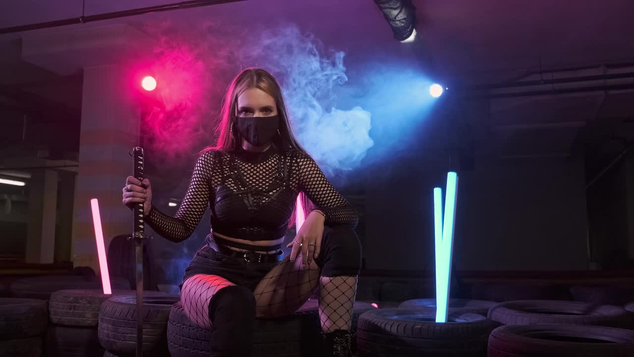戴着武士刀和面具的女孩坐在有烟雾和霓虹灯的汽车轮胎上视频下载