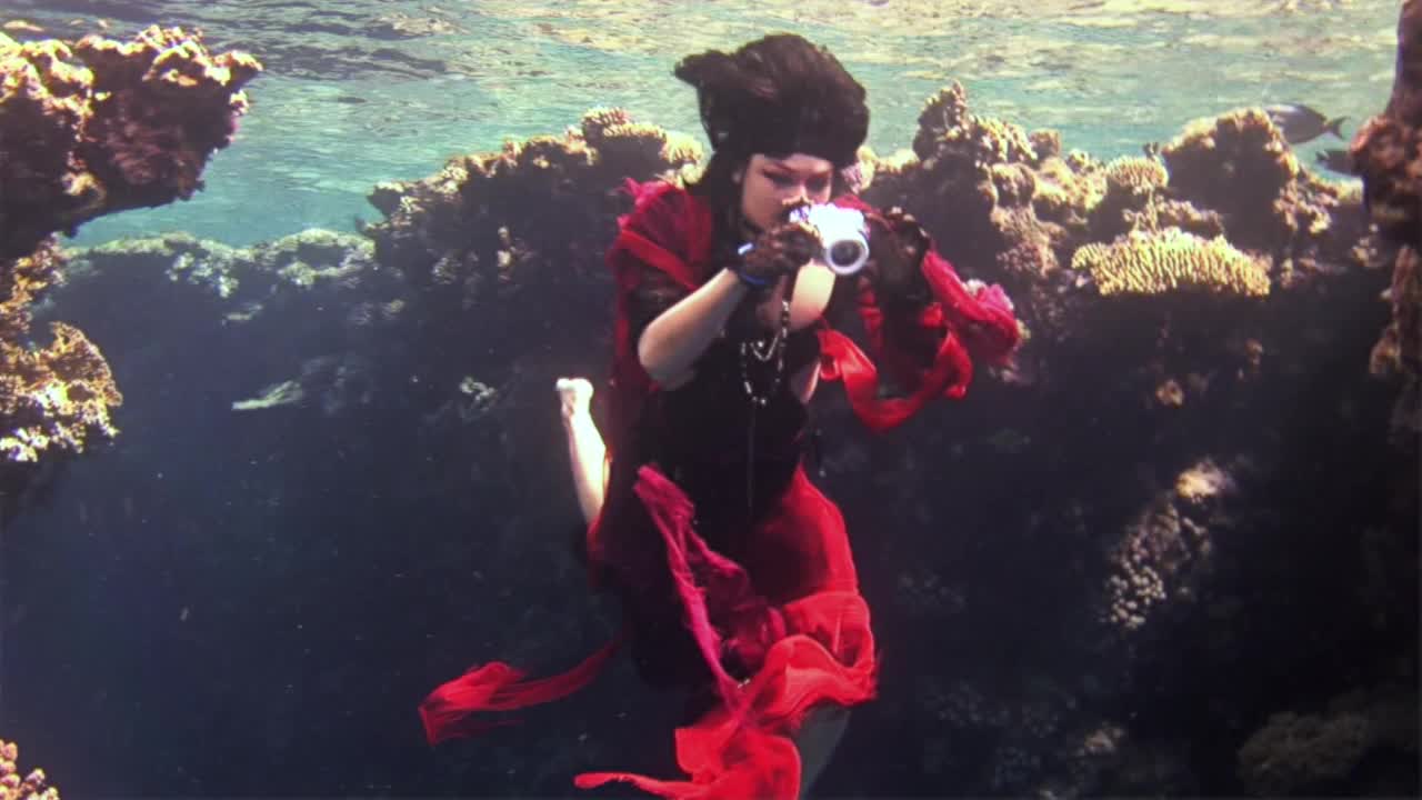 年轻的水下模型自由潜水在红海穿着红色的衣服在相机上拍照。视频下载
