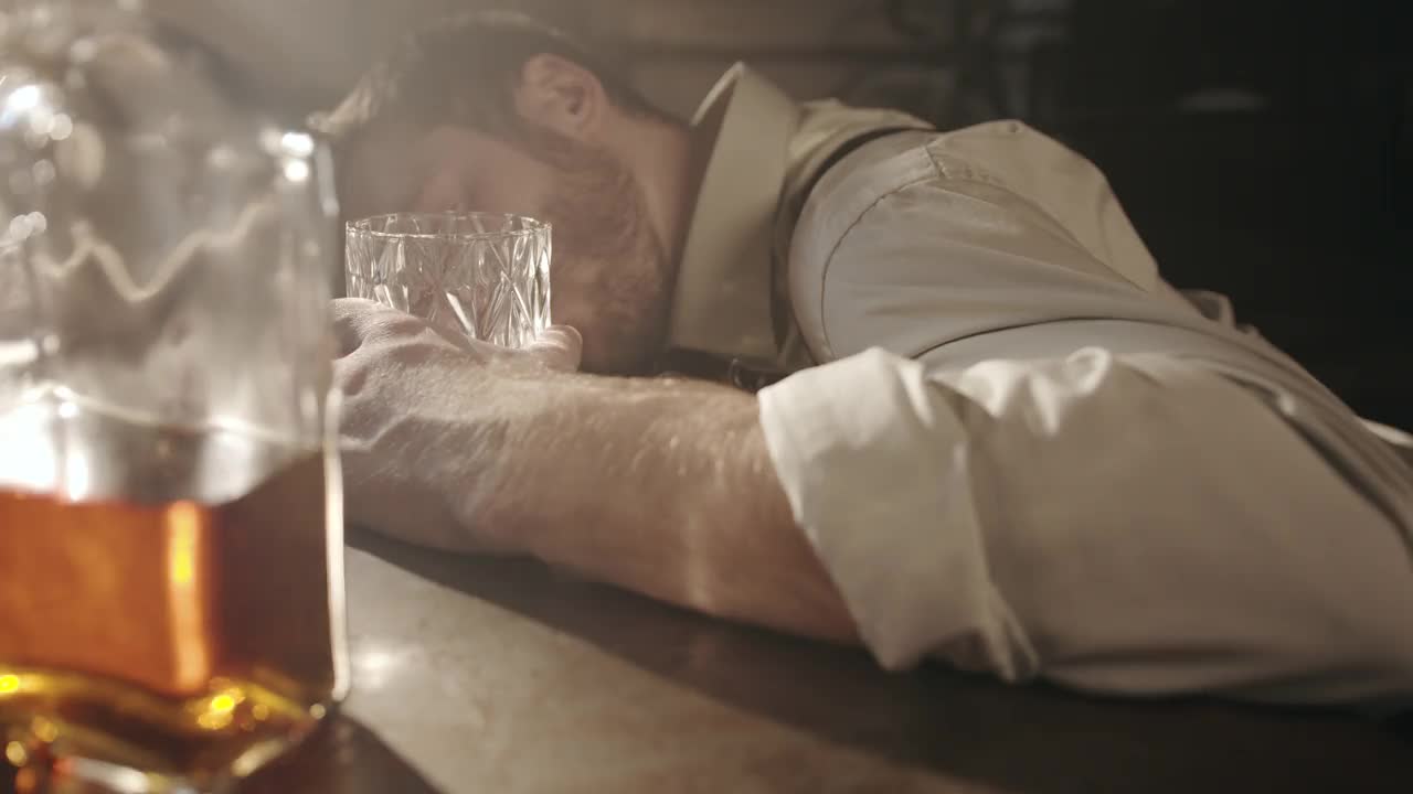 在一家昏暗的酒吧里，一个醉醺醺、疲惫不堪的男人躺在一张桌子上睡觉，手里拿着一杯白兰地，面前的桌子上放着一瓶喝得半醉的酒视频下载
