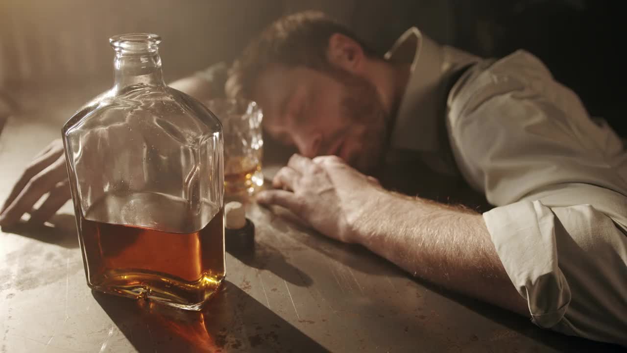 在黑暗的酒吧里，一个穿着衬衫的醉汉在桌子上睡着了，他醒了过来，从手里拿着的杯子里呷了一口威士忌，又睡着了视频下载