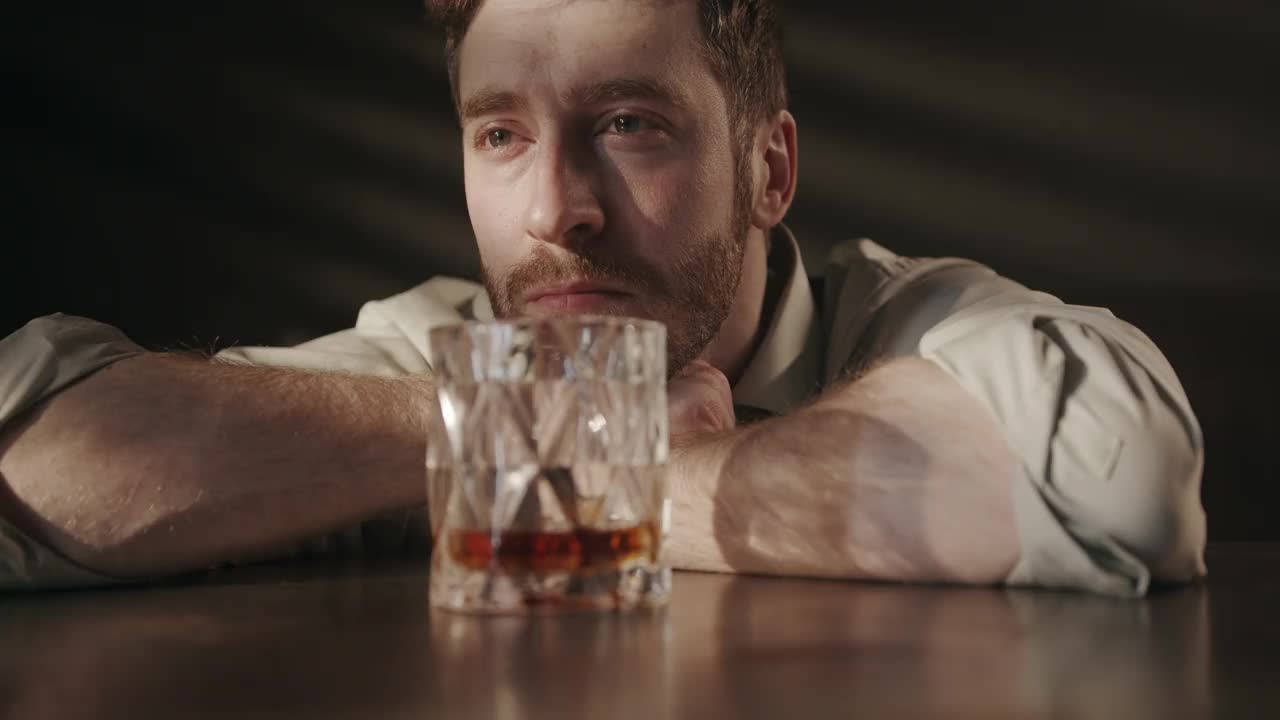 一个留着胡子的年轻醉汉双手托着头，悲伤地看着面前的那杯威士忌，思索着自己的人生视频下载
