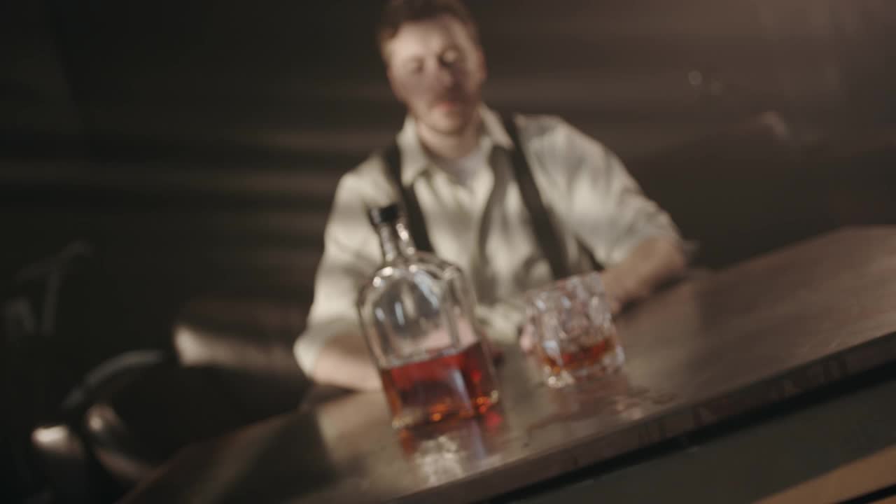 在一间黑暗的房间里，一个喝酒的人坐在桌子旁，面前放着一瓶威士忌和一个杯子，然后镜头对准了他的脸，向我们展示了他是如何喝的视频下载