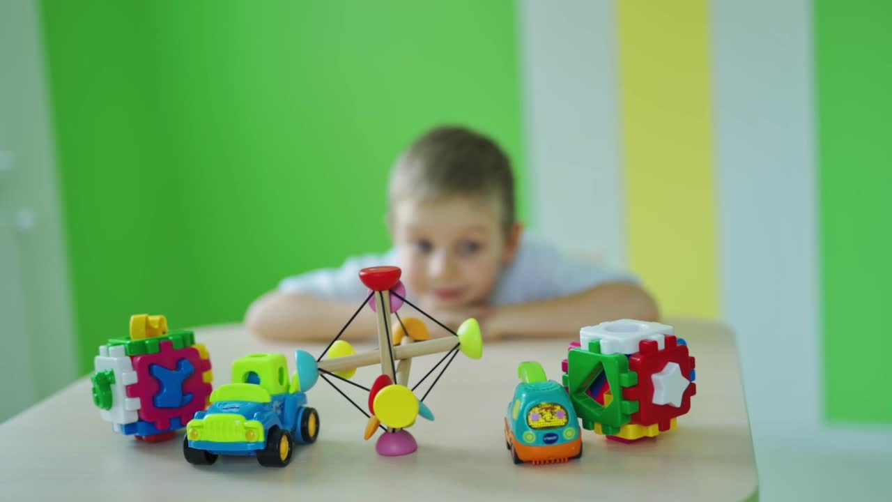 快乐的小男孩和玩具。明亮的玩具在桌子上。可爱的孩子是捕捉所有的塑料玩具在彩色的房间背景。缓慢的运动。视频下载