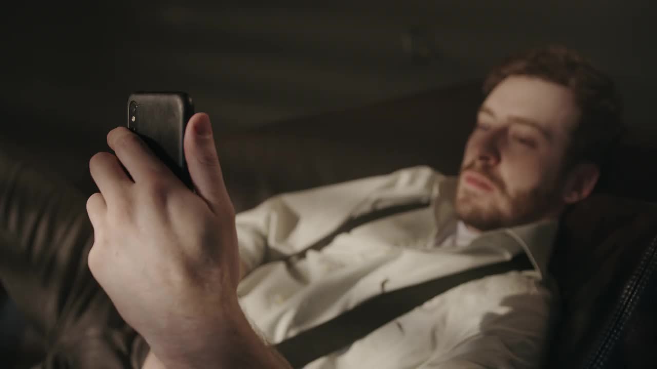 一位喝醉酒的经理下班后躺在沙发上，一边喝着威士忌一边看手机视频素材