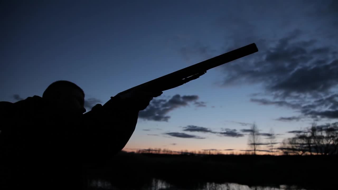 一个拿着枪的猎人的剪影。一个猎人的剪影与枪在日落的背景。一个男人拿着枪，背景是傍晚的天空和河上的狩猎视频下载