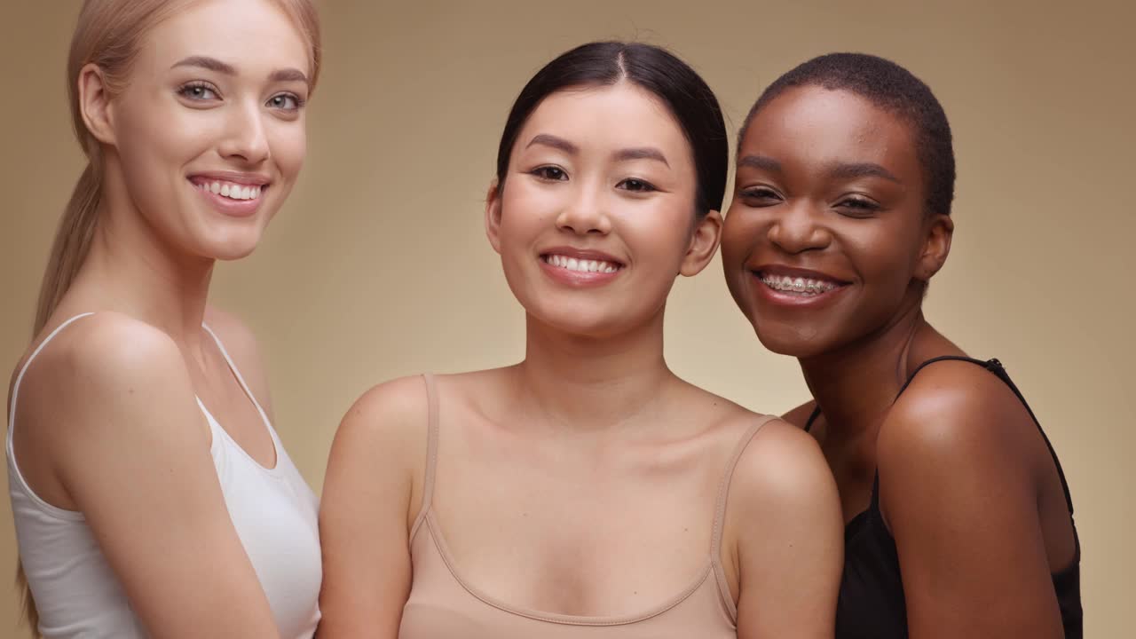 工作室的肖像三个快乐的多民族妇女笑到相机，享受友谊，拥抱在米色的背景视频下载