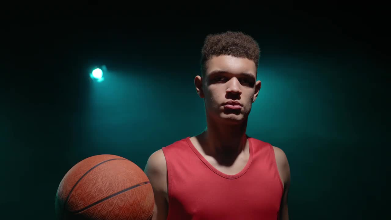 一个年轻的篮球运动员看着镜头把球抛起来的肖像。一名运动员在烟雾和蓝色灯光背景下拍摄。关闭了。慢动作准备59.94fps视频下载
