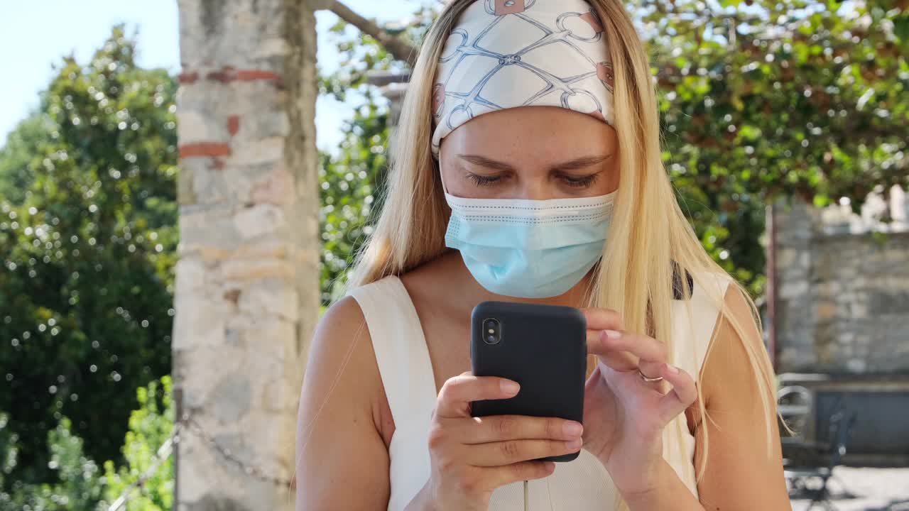 美丽的年轻女子金发博主在保护医疗面具看智能手机屏幕，并做网上购物的销售。社交媒体和技术概念。视频素材