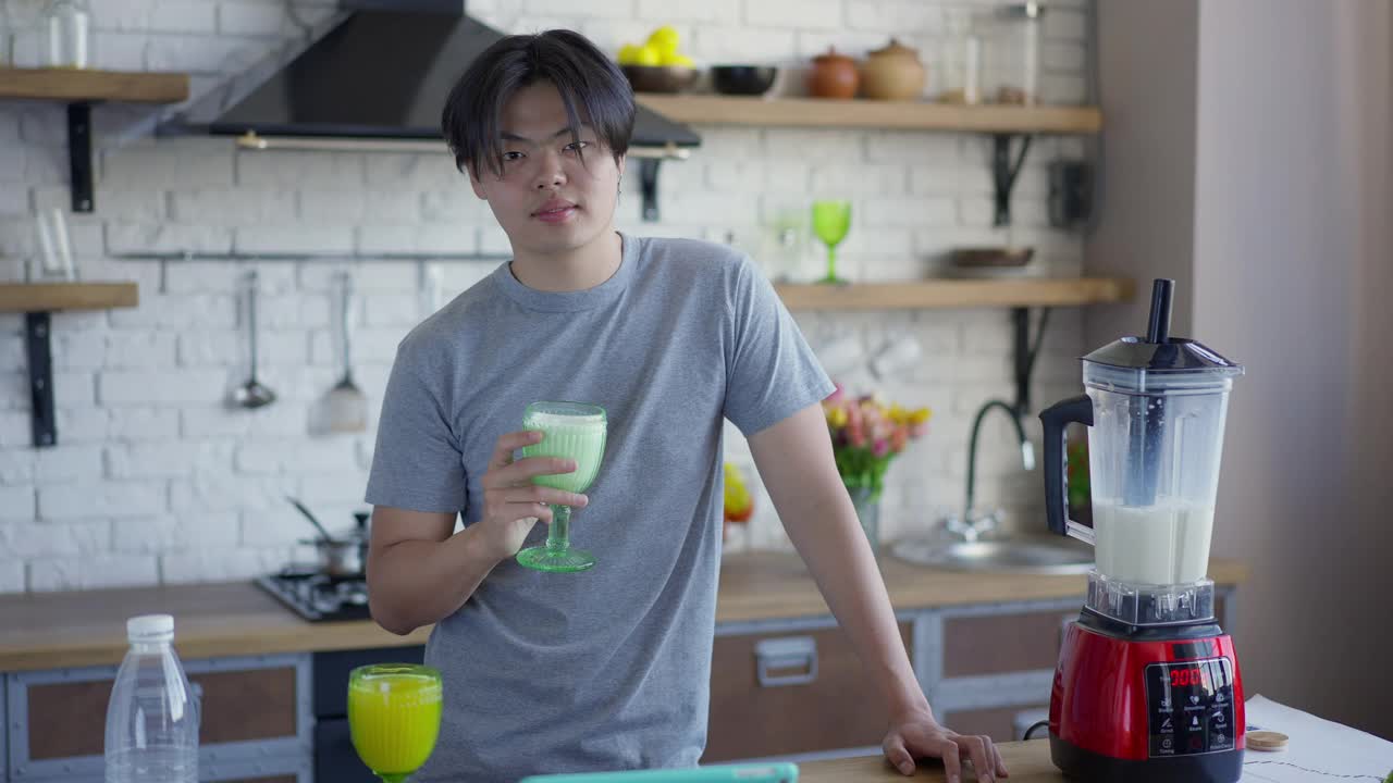 年轻快乐的亚洲男人闻着美味的牛奶鸡尾酒在杯子里微笑看着镜头。积极英俊的千禧摆姿势与健康饮料在厨房在家。生活方式和营养。视频素材