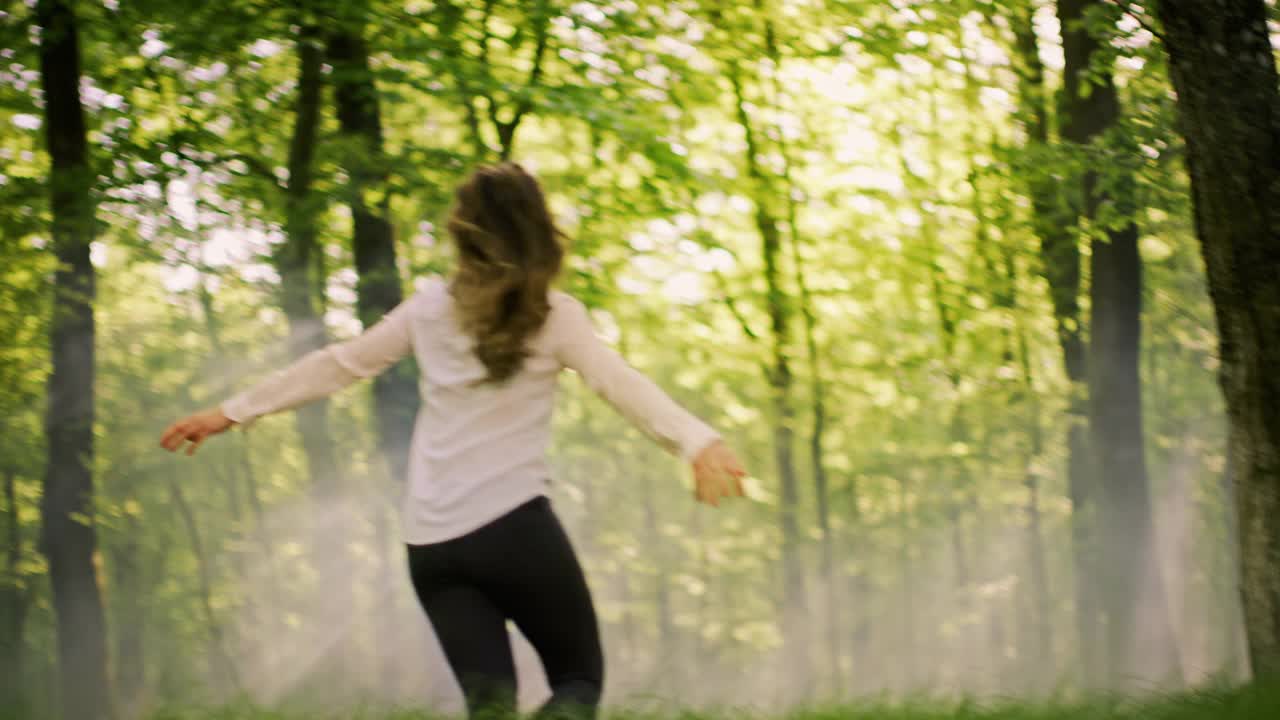 在阳光明媚的森林里，一个女人伸开双臂跑在一片绿色的空地上视频下载