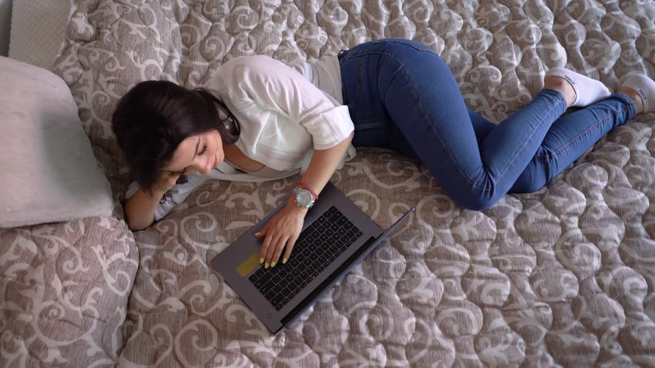 快乐的年轻女子躺在床上，在家里使用笔记本电脑视频下载