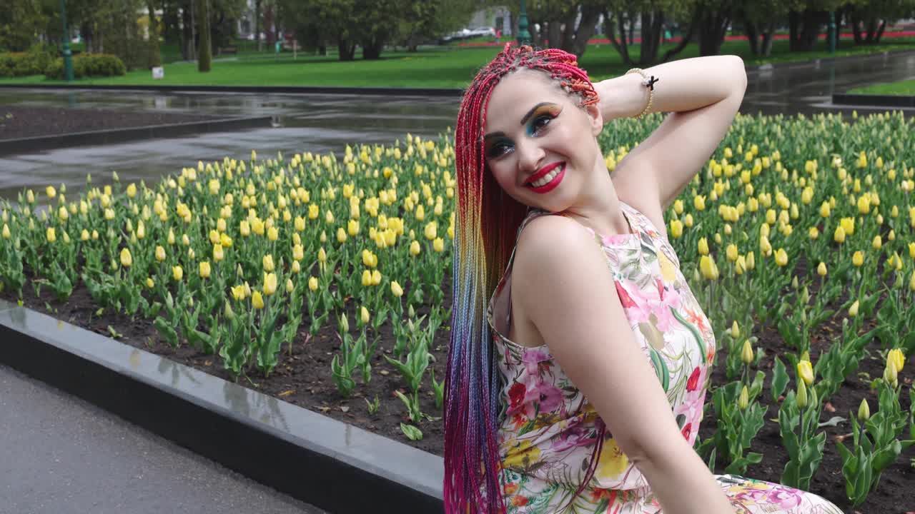 在温暖的天气里，一个化着明亮妆容、梳着非洲彩虹辫的女孩正在盛开的公园里享受春天的到来视频下载