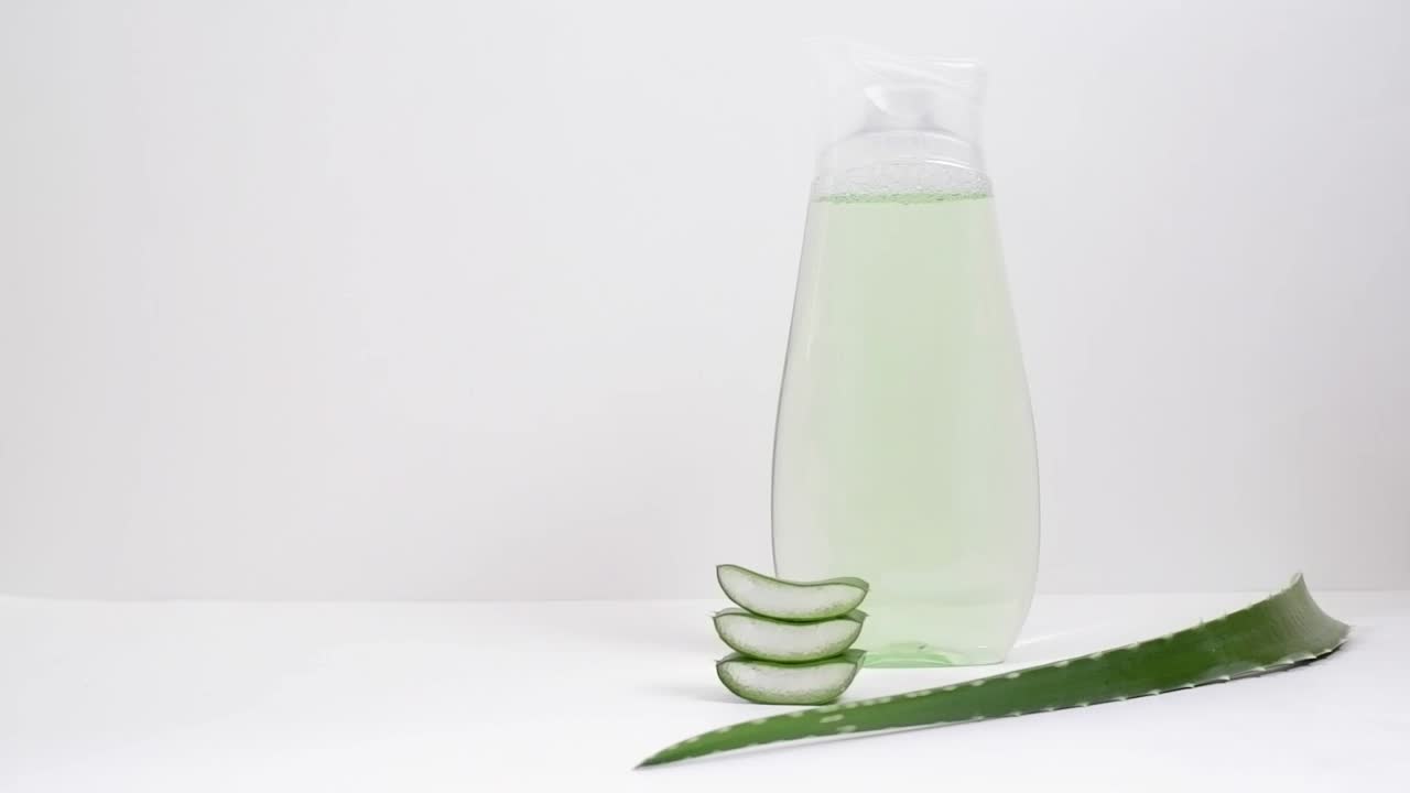 女士的手摇着一个透明的瓶子，瓶子上有绿色的乳液或补品，背景是白色的芦荟叶。水疗概念，家庭美容。视频下载