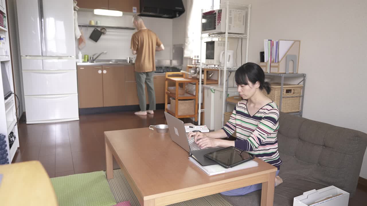女人在用笔记本电脑工作，而男人在厨房煮咖啡。视频下载