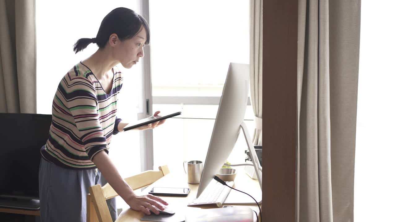 中年妇女在家里工作，用着窗边的平板电脑和电脑。视频下载