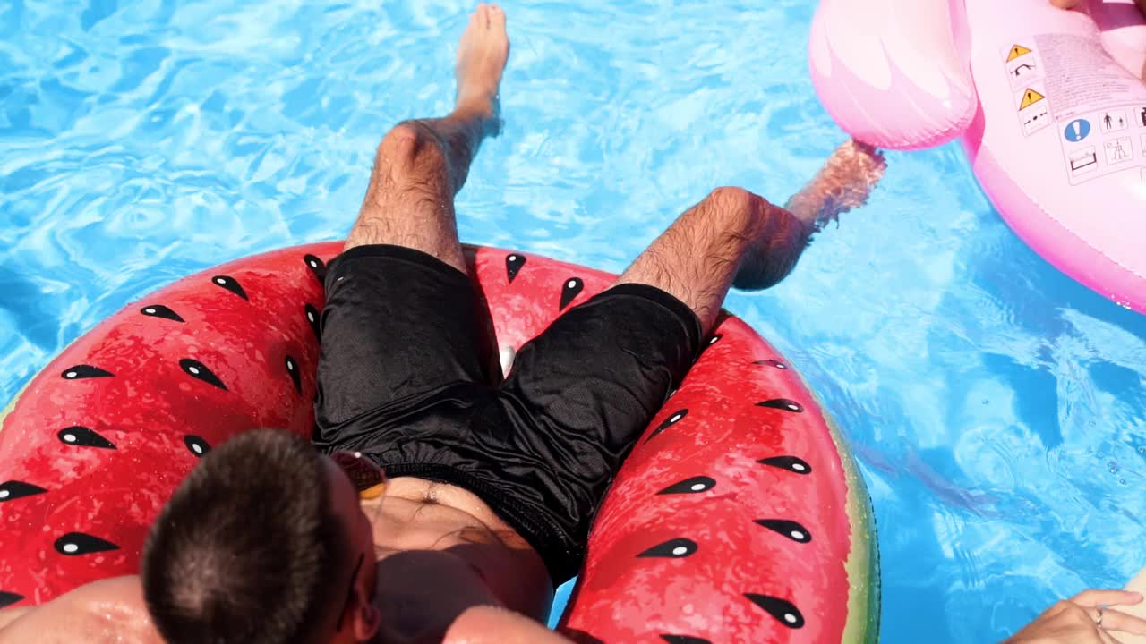 穿着短裤的男人放松在漂浮的西瓜水环上。朋友们在私人别墅的游泳池里乘凉。阳光明媚的日子，年轻人在豪华度假村的派对上放松。慢动作视频下载