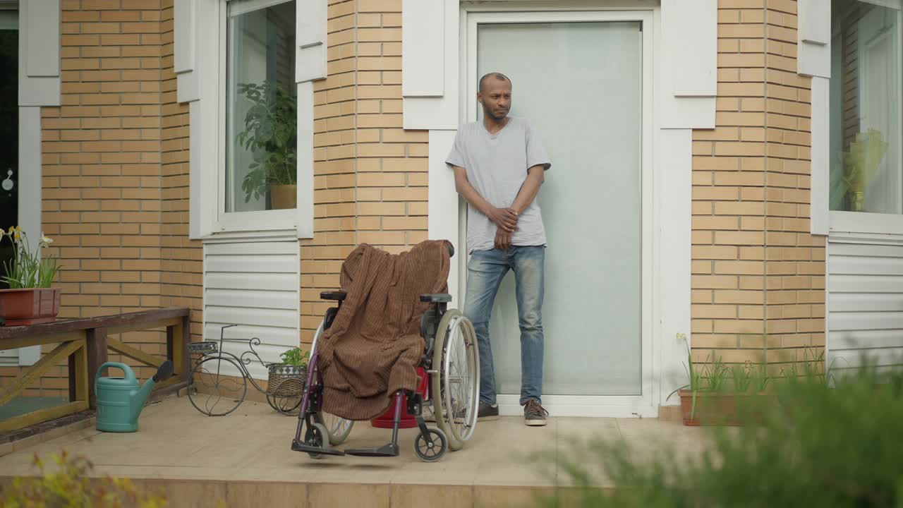 英俊的非裔美国人站在门廊上坐着轮椅等着的广角肖像。在家中或养老院照顾残疾人的成年护士或亲属。视频下载