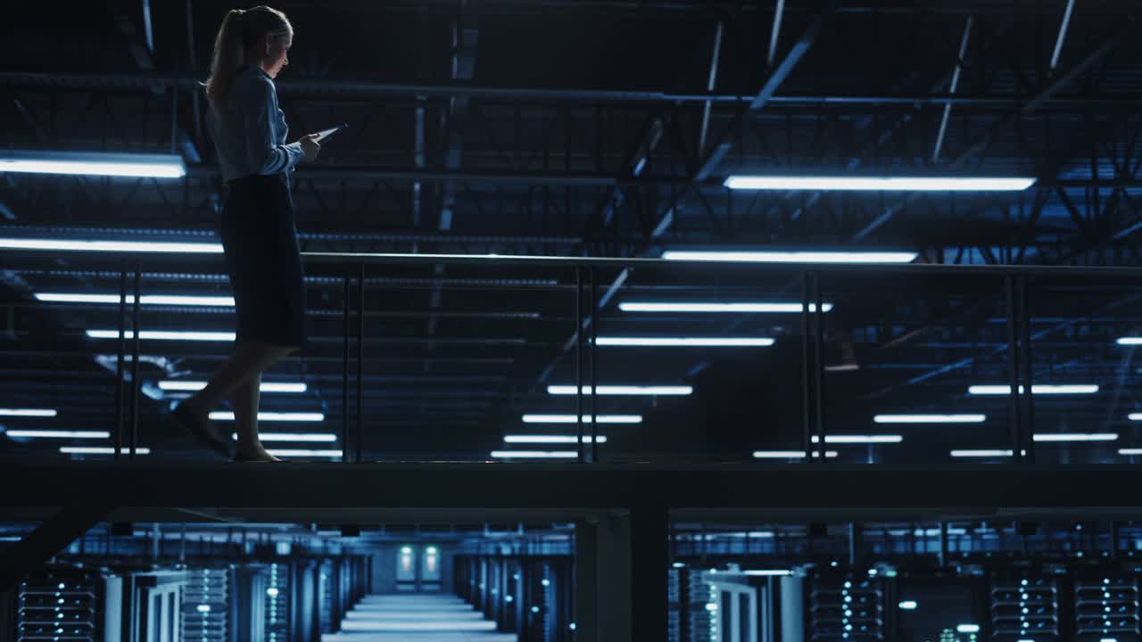 女IT专家在数据中心使用平板电脑，走在俯瞰巨大服务器农场云计算设施的桥上。成功的女商人，电子商务企业家。广角镜头视频下载